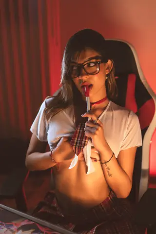 Young woman with glasses in a white shirt and plaid skirt sitting in a gaming chair under warm red lighting.