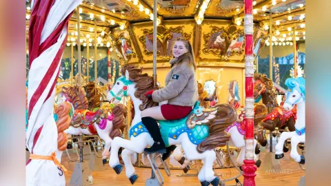 Ada — Smiling young woman in a cozy sweater riding a white carousel horse at a bright, festive carnival.