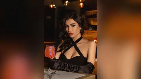 Elegant woman in a black halter dress and gloves holding a cocktail in a moody, dimly lit lounge.