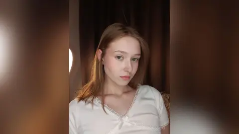 Young woman with long brown hair wearing a white lace blouse in a warm, dimly lit indoor setting.