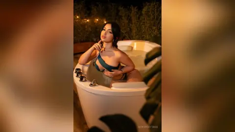 VickyPortman poses in a white bathtub filled with water, wearing a dark green bikini top and looking toward the camera.