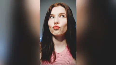 Young woman with long dark hair in a pink mesh top making a playful pouty face against a neutral background.