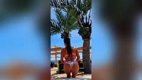 Woman in pink bikini sitting on a sunny tropical beach under palm trees near straw umbrellas.