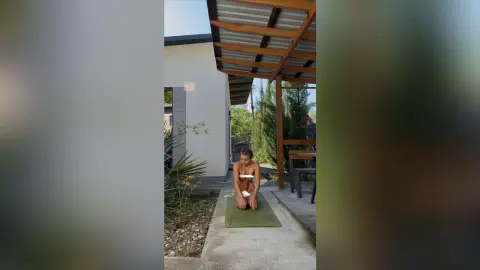 Young woman in white bikini practicing yoga on a mat in a sunny outdoor garden patio setting.