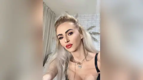 Blonde woman in a black top wearing a gold eye necklace posing with a glamorous look indoors.