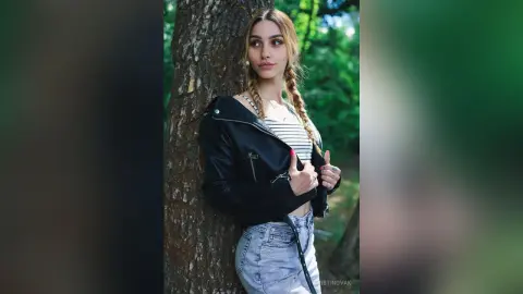 Young woman with blonde braids in a black leather jacket and denim jeans leaning against a tree in a lush forest.