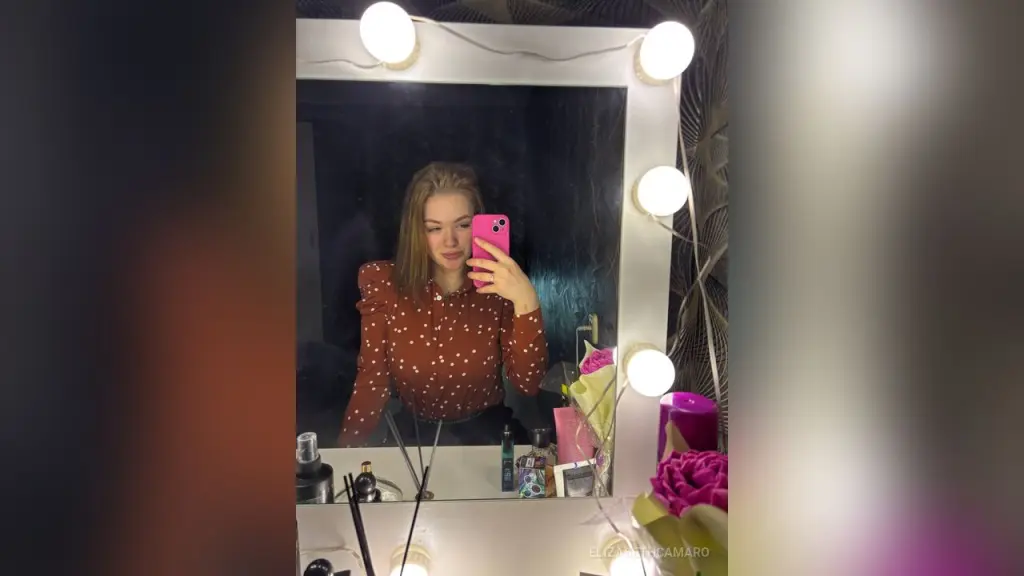 ElizabethCamaro poses for a mirror selfie wearing a pink fuzzy sweater, showing off her blonde hair and bright blue fingernails against a textured grey wall.