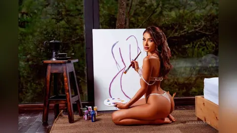 ArianaRoux kneels on a mat while holding a paintbrush and palette next to a canvas, showing her bare back and buttocks.