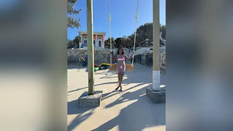NatyGlow sits on a wooden swing on a sandy beach, wearing a pink bikini and posing with her hands on the ropes.