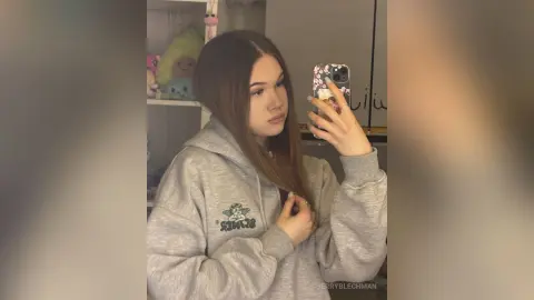 Young woman in a grey oversized hoodie taking a cozy mirror selfie in a bedroom setting.