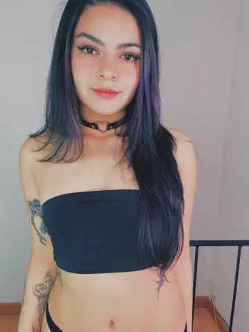 Young woman with long dark hair wearing a black tube top and choker in a minimalist indoor setting.