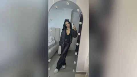 Stylish woman in a black leather jacket and trousers posing for a mirror selfie in a modern bedroom.