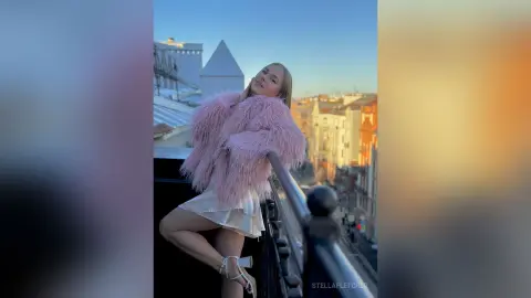 StellaFletcher poses on a balcony wearing a fluffy pink fur coat and a white mini skirt.