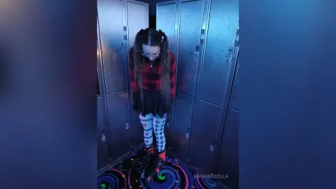 Goth girl in red plaid shirt and patterned tights standing in a blue lit locker room hallway.