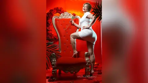 AnjaliZaharaz poses in a tight white dress while standing next to a large red velvet chair against a bright red background.