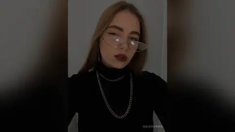 Young woman with glasses and silver chain necklace wearing a black turtleneck in a moody indoor setting.