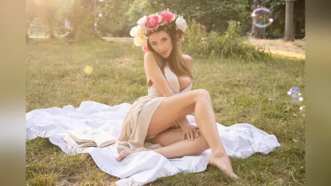 Young woman in a beige dress and flower crown sitting on a white blanket in a sunny meadow.