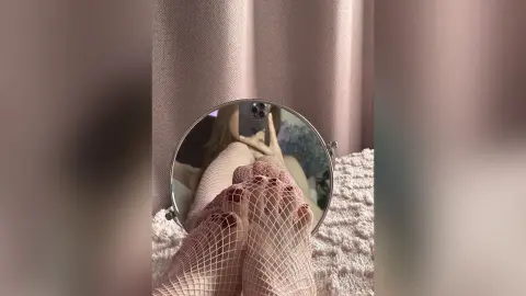 Woman in white fishnet tights relaxing on a cozy textured bed with a circular mirror reflection.