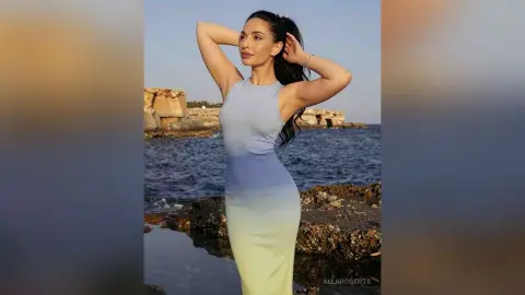 Elegant woman in a blue ombre maxi dress posing by the rocky ocean coast at sunset.