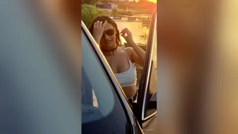 Stylish woman in sunglasses and white bikini top posing by a car at a lake during a golden hour sunset.
