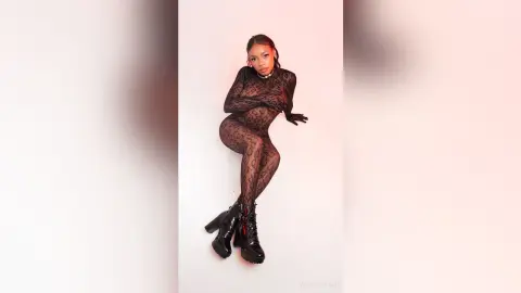 Stylish woman in a black lace bodysuit and chunky boots posing against a pink studio background.