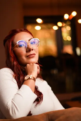 Red-haired woman in a white sweater wearing glasses, smiling thoughtfully in a warm, dimly lit cafe setting.