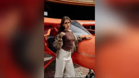 Stylish woman in a brown top and plaid cardigan posing confidently in front of a vintage orange airplane.