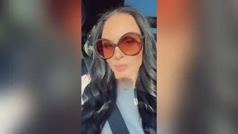 Smiling woman with long dark hair wearing oversized sunglasses in bright outdoor sunlight.
