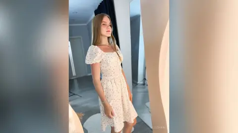 LiliLohan stands in a white floral print dress with short puff sleeves against a light-colored wall.