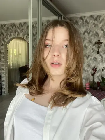 Young woman with long brown hair wearing a white shirt in a bright, cozy room.