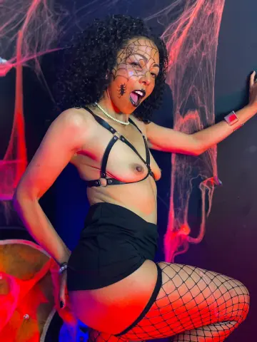 Curvy woman in black lingerie and fishnets posing against spooky spiderweb decor in moody red lighting.