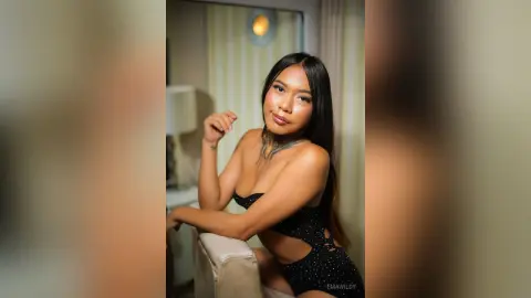 EmaWildy poses in a black cut-out bodysuit with her long dark hair down, leaning against a white chair.