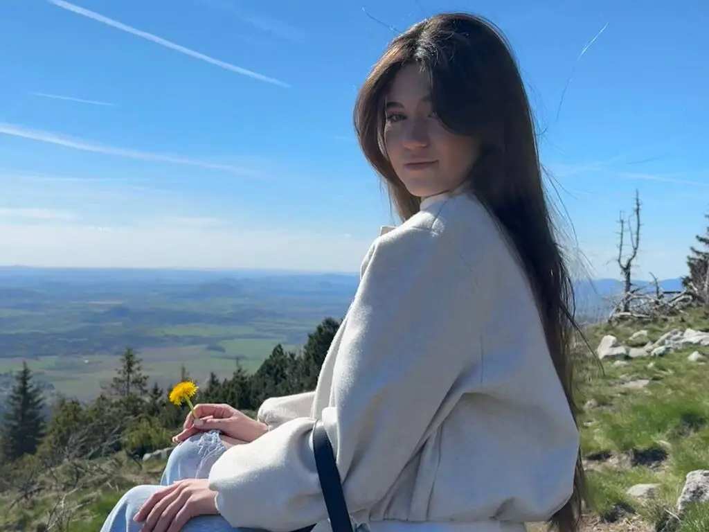 IvannaBloss sits outdoors in a white sweater and jeans, looking back over her shoulder with long brown hair against a blue sky.