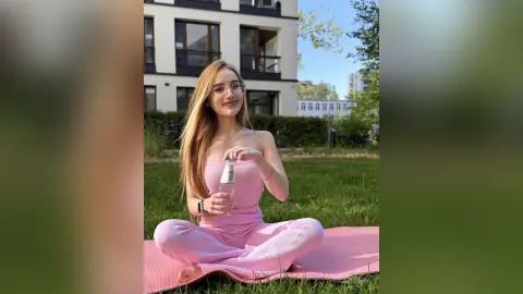 Smiling young woman in pink ribbed loungewear sitting on a yoga mat in a sunny outdoor park setting.