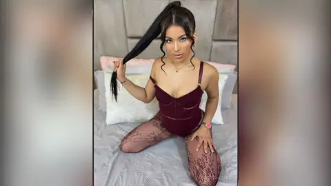 VictoriaCapri poses on a bed wearing a maroon bodysuit and lace tights, pulling back her long dark hair.
