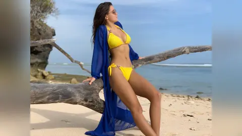 Woman in yellow bikini and blue cover-up relaxing on a sunny beach by driftwood.