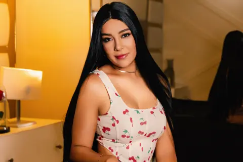 Young woman with long black hair wearing a white cherry print top in a warm, cozy indoor setting.