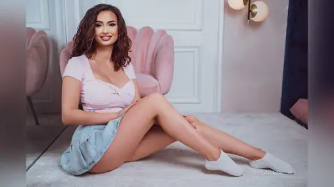 Smiling woman in a pink top and denim skirt sitting on a white rug in a stylish pink room.