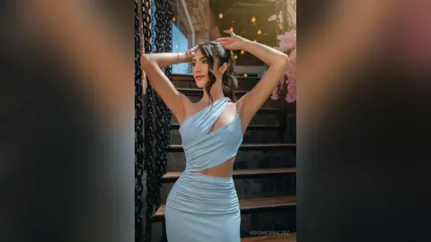 Elegant woman in a light blue one-shoulder cocktail dress posing confidently on outdoor stairs at night.