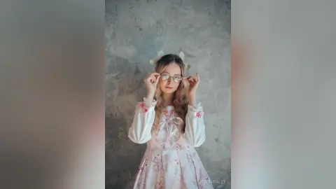 Young woman in a vintage floral white dress and glasses posing against a textured grey wall in a soft aesthetic.