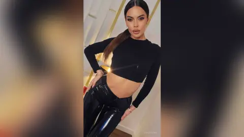 Glamorous woman in a black long-sleeve crop top and shiny black leather pants posing in a modern interior.