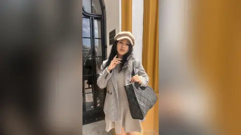 Stylish woman in a grey patterned coat and beige cap posing outside a building with a black designer tote bag.