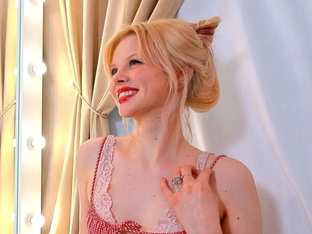 Smiling blonde woman in a red gingham lace camisole relaxing in a bright, cozy indoor setting.