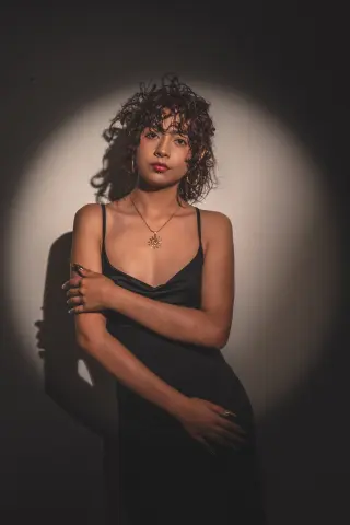 Young woman in a black slip dress and sun pendant posing under dramatic spotlight lighting.