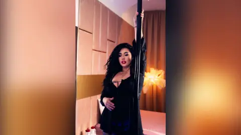 AdiraMaeven poses with a black pole, wearing a black top and showing her long, dark wavy hair in a dimly lit room.