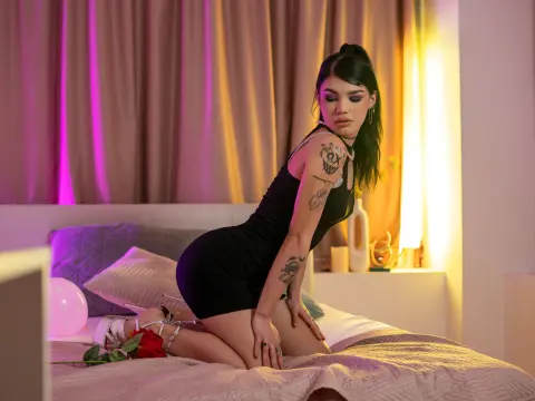 Tattooed woman in a black mini dress sitting on a bed in a moody, warm-lit bedroom setting.