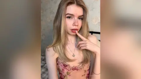 UnataMigdel is a blonde woman wearing a floral lace top and a silver heart necklace, posing with her finger to her lips.