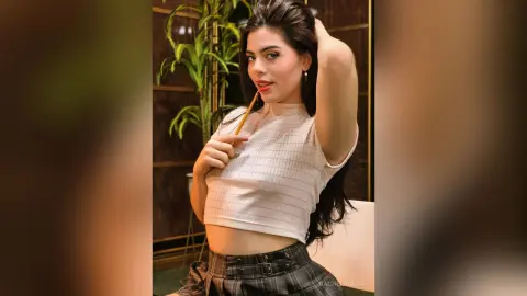 Young woman in a white ribbed crop top and plaid skirt posing confidently in a modern indoor setting.