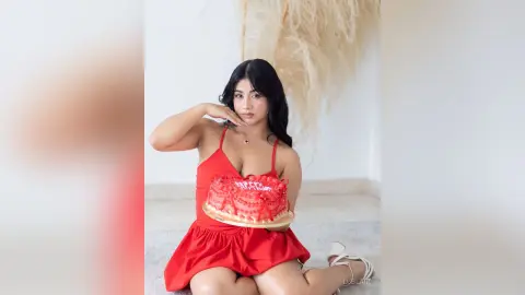 Young woman in a red summer dress holding a red cake in a bright, minimalist indoor setting.