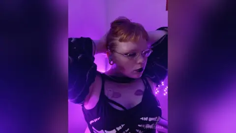 Redheaded woman in black tank top posing under moody purple lighting in a dimly lit room.